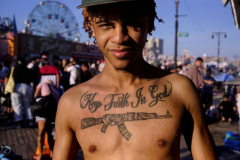 Reyes & His Tattoo - Reyes waits to wash the sand off his feet while on the boardwalk in the summer resort town of Coney Island located in Brooklyn, New York. America has a deadly fascination with guns. Over 47 thousand people were killed by firearms in 2022 and gun shots have taken over from car accidents as the number one killer of children. There has been an increase in mass shootings, over 600, in 2022 which is nearly double the number just 5 years ago.. Saturday 07 23 2022  Brooklyn, NY.