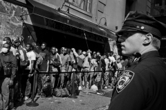 July 31, 2023 Migrant men line up outside the Roosevelt Hotel in New York City. Migrants are allowed to leave but they stay to keep their place in line, to get food and there was confusion and language barrier. 

Title: Migrants Camping in New York City Bring a Crisis into Focus: Near the bustling Grand Central Terminal in Midtown Manhattan, tired migrants waited on the sidewalks outside a former renowned hotel, once an icon of Madison Avenue. The Roosevelt Hotel is now dubbed the new Ellis Island, the latest gateway for NYC immigrants. It exemplified the city's migrant crisis at a critical juncture.

A year ago, the migrant crisis in NYC peaked as GOP governors from Texas and Florida bused migrants to the city in 2022, seeking to attribute the situation to Biden's immigration policies. Since 2022, more than 150,000 migrants have come to NYC. For the first time, migrant men were left on the sidewalk during a NYC heatwave as the city gave priority to women and children. The mayor stated, "the city is running out of housing spaces."  They remained there for five days in August 2023.

As the city works to amend Title 42, a 1970s "Right to Shelter Law" meant for emergency homeless housing, NYC grapples with a daily cost of $400 per migrant to house and shelter the migrants.  This will amount to $12 billion for the city over the next three years.
