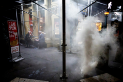 NEW YORK, NY USA - JUNE 10, 2025 - Near Federal Plaza in Lower Manhattan, two older men linger over slices of pizza as smoke drifts through the street. Blocks down the street the conflict continues as protesters and police collide over a van carrying arrested immigrants to a detention facility in New Jersey. As tension fills the air, New York City continues its quiet, unbothered rhythm.