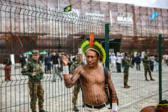 An Amazonian Indigenous man wearing traditional body paint and a feathered headdress stands at a security fence as police patrol outside the COP30 venue, where security was heightened after earlier demonstrations.