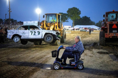 AUGUSTA, NJ, USA - AUGUST 3, 2025 - A heavy loder pauses, taking a wrecked car out of the arena as an elderly man drives by on his scooter. The first night of the 2025 Demolition Derby during the NJ State Fair held at the Sussex County Fairgrounds. The Derby is the biggest event at the fair and families pass the spectacle down through generations, and a win earns drivers a year’s worth of bragging rights.